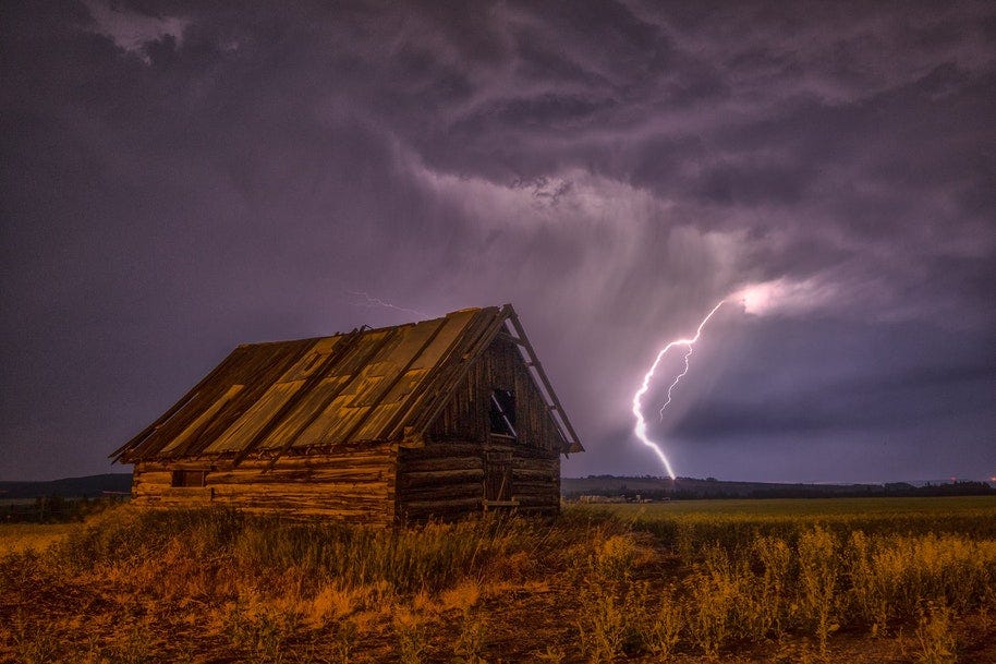 Wooden hut in open ground with lightning beyond.